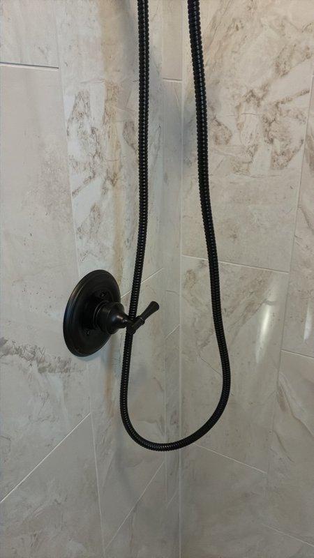 Completed fixture installation and finishing work for bathroom shower system, including installation of matte black dual-head shower configuration with overhead rain showerhead and handheld shower attachment with flexible hose and wall-mounted holder on marble-look tile surround. All fixtures installed, tested, and confirmed operational per manufacturer specifications.