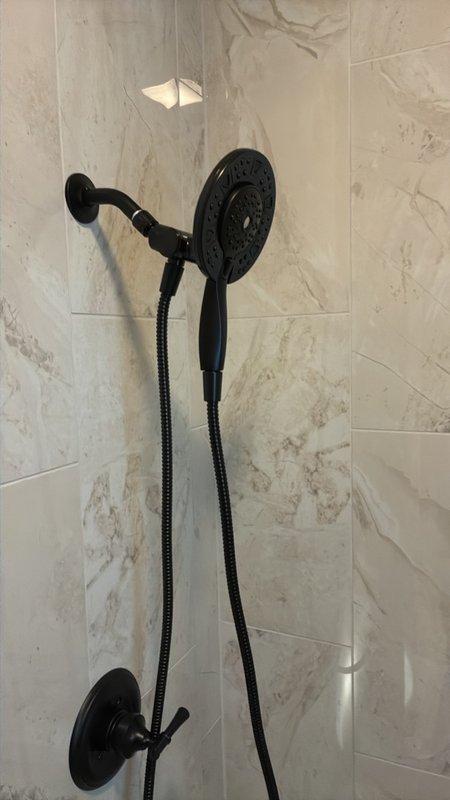Completed fixture installation and finishing work for bathroom shower system, including installation of matte black dual-head shower configuration with overhead rain showerhead and handheld shower attachment with flexible hose and wall-mounted holder on marble-look tile surround. All fixtures installed, tested, and confirmed operational per manufacturer specifications.