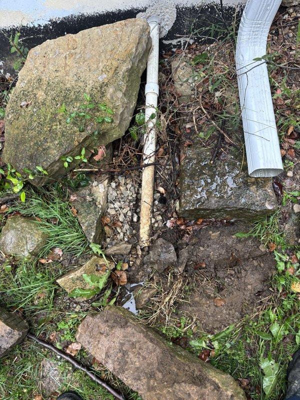 Inspected the existing sump pit and pump system to verify operation, noting the pit cover in place and the power cord properly plugged into the outlet. Observed silt and mineral staining along the basement wall base and floor indicating prior water intrusion, and checked the exterior discharge line and downspout termination, which showed staining and saturated, muddy soil at the discharge point.