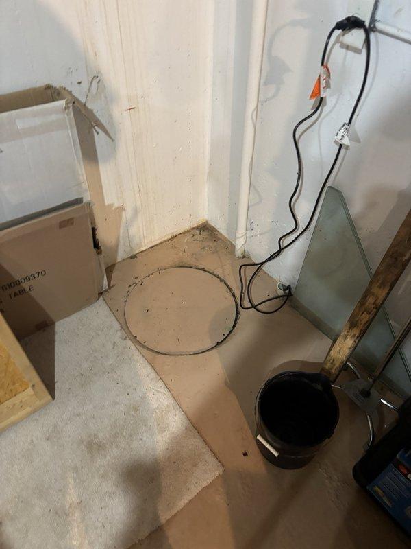 Inspected the existing sump pit and pump system to verify operation, noting the pit cover in place and the power cord properly plugged into the outlet. Observed silt and mineral staining along the basement wall base and floor indicating prior water intrusion, and checked the exterior discharge line and downspout termination, which showed staining and saturated, muddy soil at the discharge point.