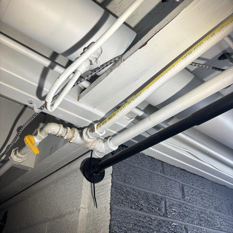 Assessed the gas supply for the new range, where a yellow corrugated flexible gas connector runs overhead through the joist space to a shutoff valve with threaded black iron fittings showing light surface corrosion and rust staining at the joints. Evaluated replacement of the flexible connector with a rigid gas pipe run and reviewed the refrigerator water supply, where the existing T-valve would need to be replaced with a proper dedicated shutoff for the ice maker/water line hookup. Documented existing conditions with photos and prepared the scope of work for the fixture installation estimate.