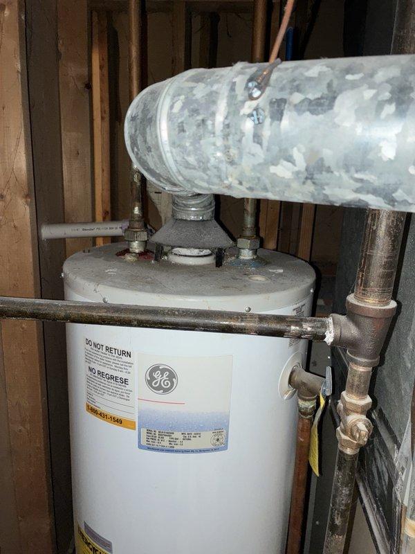 Performed an on-site evaluation of a no-hot-water complaint on a GE tank-style water heater installed in an unfinished framed space with a galvanized draft hood and flue vent. Inspected the unit and surrounding supply piping, noting tarnished and oxidized copper lines, a brass tee with white residue at the solder joint, and heavy dust and grime on the tank. Documented the installation with photographs and prepared a replacement/repair estimate for the customer's review.