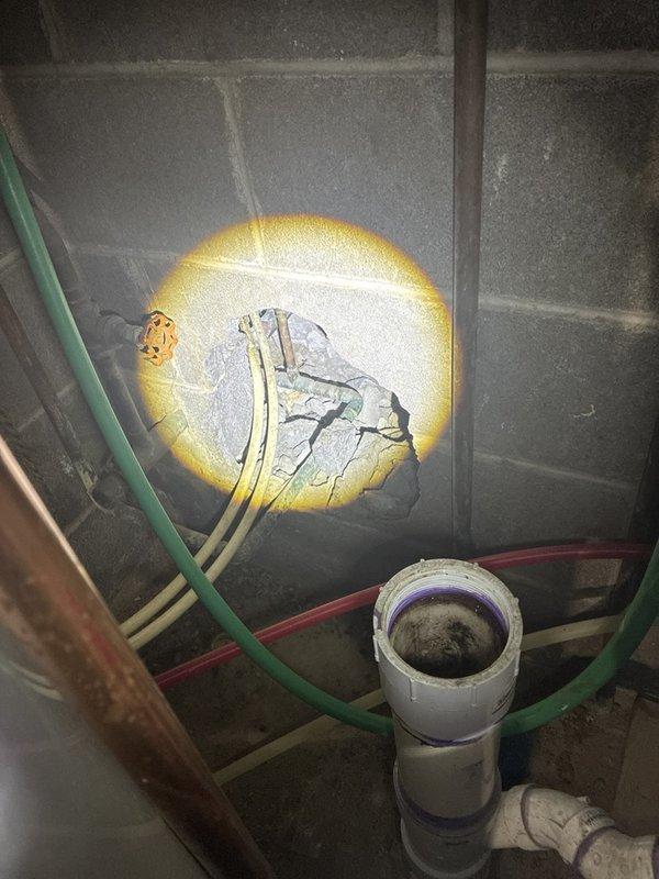 Performed a plumbing estimate to evaluate and scope repairs for reported leaks. Inspected supply and drain piping in the basement/crawlspace area, documenting a broken-out opening in the concrete block wall with pipe penetrations and fittings, corroded piping and a valve, and a PVC drain stub with a threaded coupling that is open and contaminated with debris. Also identified a tarnished chrome pipe fitting with escutcheon at a ceiling/wall penetration where deteriorated sealant and failed surface material have created a gap, indicating a probable leak source requiring repair.