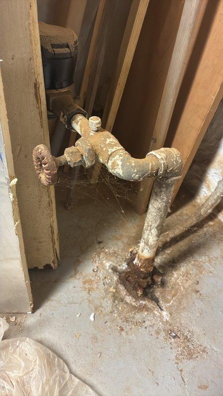 Performed an on-site assessment and estimate for boiler/water heater service, documenting the existing piping and valve assembly with photographs. Inspection revealed severe corrosion and heavy mineral scale on the vertical supply piping, elbow, and gate valve, with active rust staining and calcium deposits at the pipe base indicating long-term moisture exposure and probable seepage at the floor connection. Findings support replacement of the deteriorated piping, fittings, and isolation valve as part of the recommended scope of work.