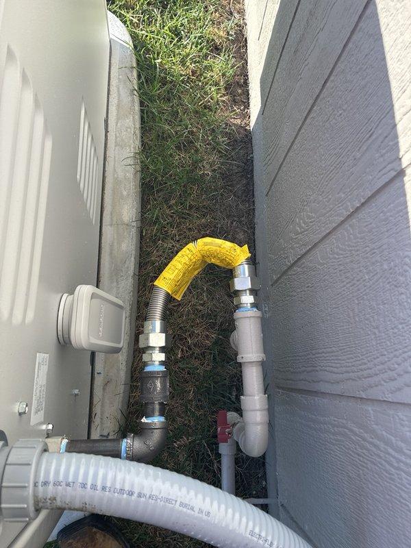 Assessed the gas piping run at the standby generator and prepared an estimate to install the manufacturer-supplied corrugated flexible gas connector between the generator's black iron outlet and the previously installed rigid gas line. The existing configuration includes a black iron nipple and elbow, a shutoff valve on the gray-painted supply pipe, and threaded fittings at both ends of the proposed connector run. Documented the existing piping and fittings with photos to support the installation scope.