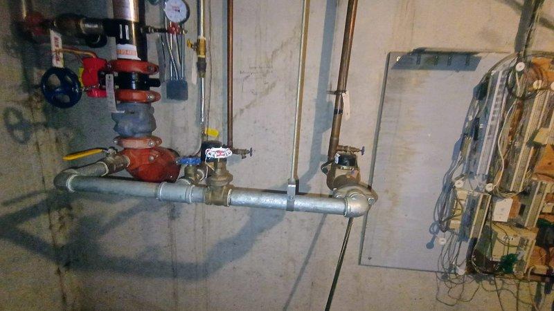 Responded to a reported water leak between the building and sprinkler system. Upon inspection, identified a compromised valve assembly on the main water line supplying the irrigation system. Visually documented the existing installation, which showed signs of water damage and mineral buildup around connection points. The brass valve with red and blue handle was tagged for replacement, as it was determined to be the source of the leak.