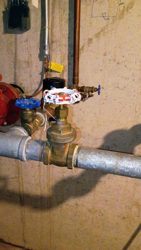 Responded to a reported water leak between the building and sprinkler system. Upon inspection, identified a compromised valve assembly on the main water line supplying the irrigation system. Visually documented the existing installation, which showed signs of water damage and mineral buildup around connection points. The brass valve with red and blue handle was tagged for replacement, as it was determined to be the source of the leak.