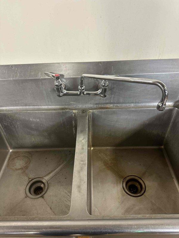 Successfully replaced faucet at commercial wash station featuring a double basin stainless steel sink. Installed new gooseneck-style faucet with dual hot/cold handles, ensuring proper connection to existing plumbing system.