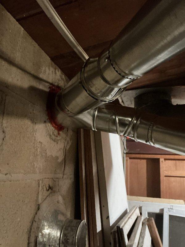 Installed new metal chimney liner through concrete block wall in basement utility area. Applied red fire-stopping sealant at wall penetration point to ensure proper sealing and fire code compliance. Connected and secured parallel galvanized ductwork runs with metal bands/clamps along ceiling area, completing the ventilation system installation.
