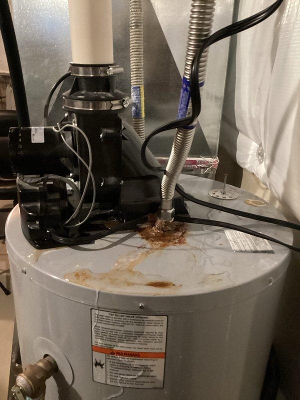 # Tanked Water Heater Diagnostic Service

Responded to customer report of water heater issue isolated to specific area, ongoing for one week. Upon arrival, conducted comprehensive diagnostic inspection of residential tanked water heater system in 6-year-old home.

Visual inspection revealed significant rust-colored staining and moisture damage on top surface of white cylindrical water heater tank, indicating active water leakage or corrosion. Brownish-orange discoloration and water stains spread across white painted metal surface, confirming ongoing moisture intrusion. Examined recirculation pump system mounted on top of tank, including black circulator unit with metal clamps securing it to white PVC piping.

Inspected all connection points where flexible metal conduit hoses extend upward from pump connections, noting blue tape wrapping on portions of conduits. Traced electrical wiring and black cables routed alongside piping system to verify proper connections. Evaluated condition of 