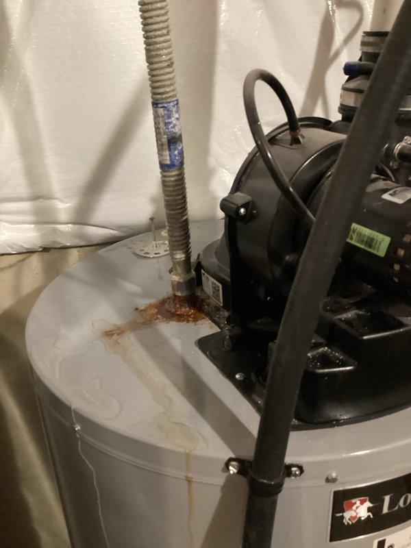 # Tanked Water Heater Diagnostic Service

Responded to customer report of water heater issue isolated to specific area, ongoing for one week. Upon arrival, conducted comprehensive diagnostic inspection of residential tanked water heater system in 6-year-old home.

Visual inspection revealed significant rust-colored staining and moisture damage on top surface of white cylindrical water heater tank, indicating active water leakage or corrosion. Brownish-orange discoloration and water stains spread across white painted metal surface, confirming ongoing moisture intrusion. Examined recirculation pump system mounted on top of tank, including black circulator unit with metal clamps securing it to white PVC piping.

Inspected all connection points where flexible metal conduit hoses extend upward from pump connections, noting blue tape wrapping on portions of conduits. Traced electrical wiring and black cables routed alongside piping system to verify proper connections. Evaluated condition of 