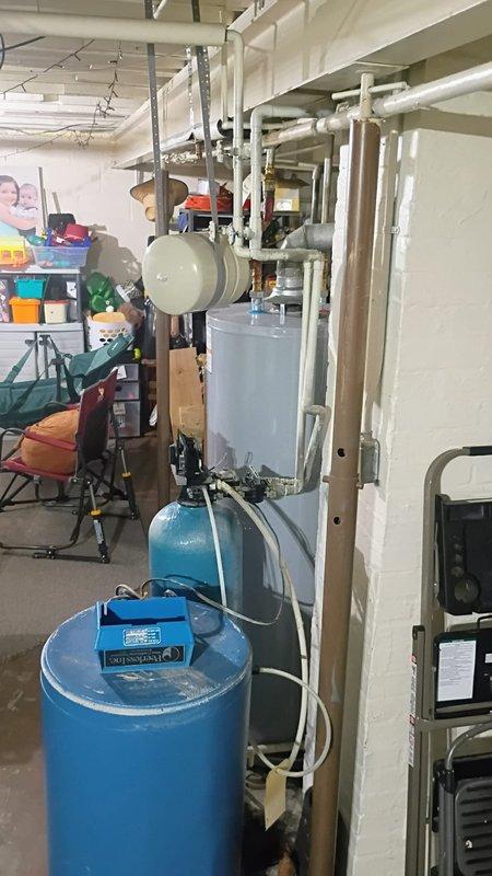Conducted on-site assessment of existing water softener system in basement mechanical room, which is over 20 years old and requires replacement. Documented current plumbing configuration including blue cylindrical water softener tank, well pressure tank, water heater, and associated PVC piping infrastructure. Noted significant water damage and flooding throughout basement utility area, with standing water covering concrete floor surface. Photographed existing equipment layout and plumbing connections to prepare comprehensive replacement estimate for new water softener installation, including necessary modifications to accommodate updated system and address water intrusion issues.