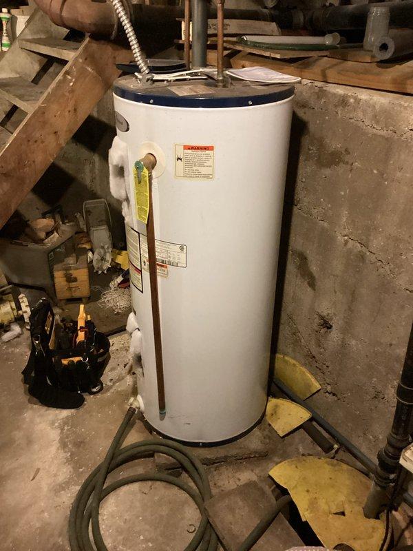 Responded to no hot water complaint and performed diagnostic inspection of tanked water heater system. Upon arrival, assessed white cylindrical tank-style water heater unit located in basement utility area and evaluated all visible components including top-mounted plumbing connections, copper supply lines, flexible metal conduit, shutoff valves, and base fittings. Documented existing conditions through photographic evidence showing water heater installation, surrounding plumbing infrastructure, and potential moisture concerns indicated by floor discoloration and staining around unit base.