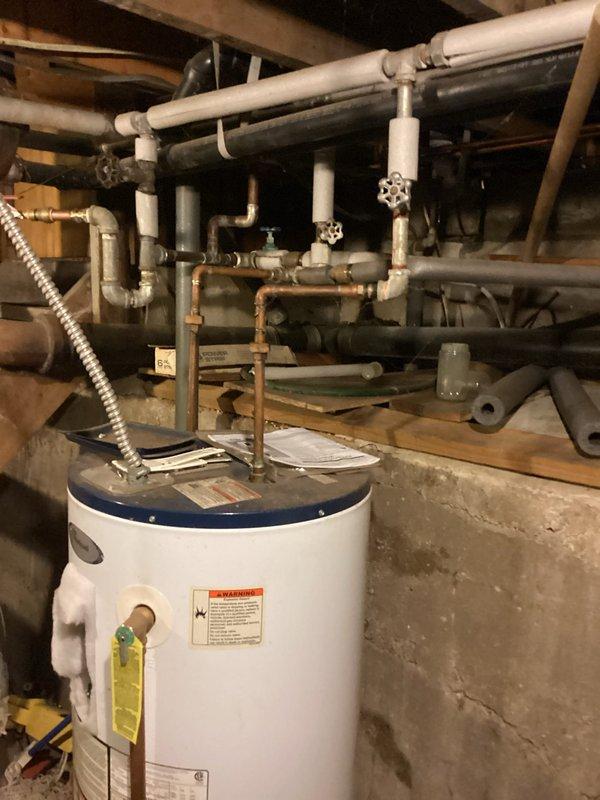 Responded to no hot water complaint and performed diagnostic inspection of tanked water heater system. Upon arrival, assessed white cylindrical tank-style water heater unit located in basement utility area and evaluated all visible components including top-mounted plumbing connections, copper supply lines, flexible metal conduit, shutoff valves, and base fittings. Documented existing conditions through photographic evidence showing water heater installation, surrounding plumbing infrastructure, and potential moisture concerns indicated by floor discoloration and staining around unit base.