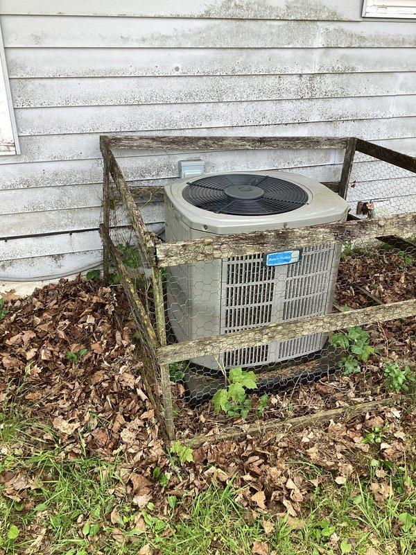 # HVAC Maintenance - Planned Single Maintenance

Completed planned air conditioning maintenance on outdoor condenser unit. Upon arrival, found unit heavily overgrown with vegetation including vines and climbing plants growing through ventilation grilles and across top surface. Surrounding wooden enclosure showed significant deterioration and decay. 

Cleared dense vegetation from condenser unit and removed accumulated leaf debris from base and surrounding area. Cleaned exterior surfaces and ventilation grilles to restore proper airflow. Inspected unit components and verified operational status. Recommended removal or repair of deteriorated wooden structure to prevent future vegetation encroachment and ensure adequate clearance for service access and airflow requirements.