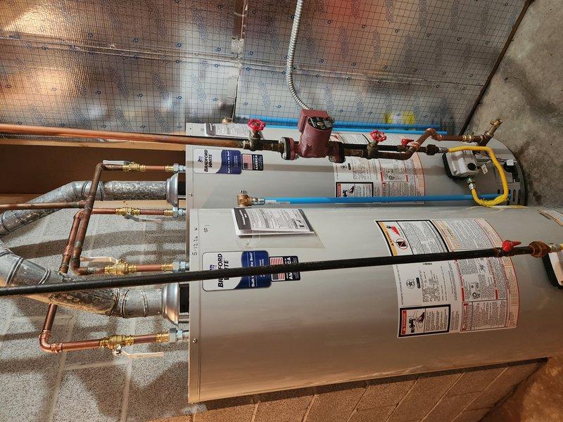 Completed installation of two Bradford White water heaters in utility room. Units were positioned side by side on tiled flooring and connected to existing plumbing infrastructure using copper piping with shutoff valves, flexible connectors, and yellow flexible gas lines for fuel supply. All connections were properly secured and tested for safe operation.