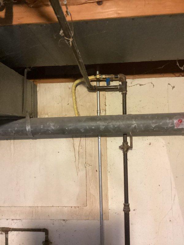 Responded to gas leak detected by gas company during meter replacement at approximately 60-year-old residential property, resulting in complete gas shutoff to home. Performed comprehensive pressure testing on central heating system and gas distribution lines to identify source of leak in aging styrene gas piping system measuring approximately one to one-and-a-half inches in diameter. Inspected exposed plumbing infrastructure in basement utility area, documenting condition of gas lines, connections, and fittings throughout the system. Evaluated yellow flexible gas connectors, metallic piping, and junction points for integrity and compliance. Photographed existing conditions showing weathered gray gas lines, multiple pipe connections, and aged infrastructure components mounted along floor joists and foundation walls.