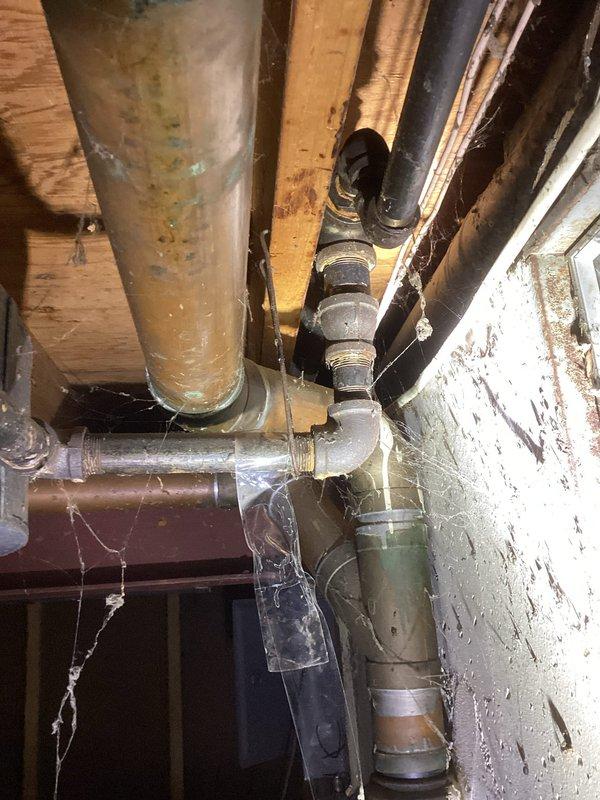 Responded to gas leak detected by gas company during meter replacement at approximately 60-year-old residential property, resulting in complete gas shutoff to home. Performed comprehensive pressure testing on central heating system and gas distribution lines to identify source of leak in aging styrene gas piping system measuring approximately one to one-and-a-half inches in diameter. Inspected exposed plumbing infrastructure in basement utility area, documenting condition of gas lines, connections, and fittings throughout the system. Evaluated yellow flexible gas connectors, metallic piping, and junction points for integrity and compliance. Photographed existing conditions showing weathered gray gas lines, multiple pipe connections, and aged infrastructure components mounted along floor joists and foundation walls.