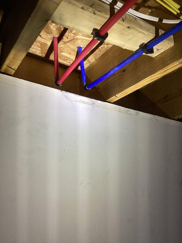 Replaced plastic water supply fittings with code-compliant metal fittings in exposed ceiling area as required by building inspector. The work involved the hot and cold water supply lines (identified by red and blue PEX tubing) running through the ceiling joists, where plastic compression fittings were removed and replaced with brass or copper transition fittings. All connections were properly secured to the wooden framing members using appropriate mounting hardware. Photographed completed installation showing the metal fittings installed at junction points, electrical box location noted for clearance purposes, and overall condition of the work area. All replaced fittings now meet current plumbing code requirements for inspection approval.