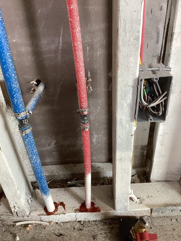 Replaced plastic water supply fittings with code-compliant metal fittings in exposed ceiling area as required by building inspector. The work involved the hot and cold water supply lines (identified by red and blue PEX tubing) running through the ceiling joists, where plastic compression fittings were removed and replaced with brass or copper transition fittings. All connections were properly secured to the wooden framing members using appropriate mounting hardware. Photographed completed installation showing the metal fittings installed at junction points, electrical box location noted for clearance purposes, and overall condition of the work area. All replaced fittings now meet current plumbing code requirements for inspection approval.