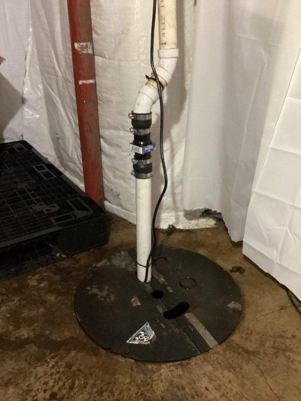 Responded to emergency flooding situation in basement caused by failed sump pump system. Upon arrival, found approximately 1 inch of standing water covering 15-foot radius around sump pit. Assessed existing sump pump installation, documented wet concrete floor conditions and active water infiltration around corroded metal pit cover. Photographed complete system including vertical PVC discharge piping with mounting straps, electrical connections, and moisture barrier installation on surrounding walls.