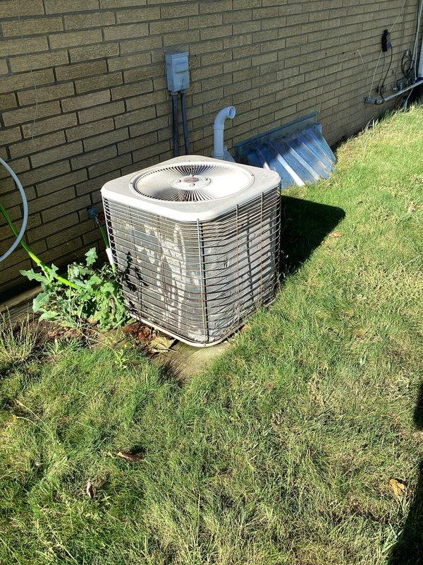 Responded to no-cool complaint with outdoor unit not running and warm air from vents. Upon arrival, found white cylindrical condenser unit positioned against exterior brick wall with airflow present at indoor registers and no ice buildup at evaporator coil. Performed visual inspection of outdoor unit, verified electrical disconnect box and refrigerant line connections, and documented condenser placement and surrounding installation components including PVC condensate drain and refrigerant line routing along foundation. System age unknown but reported as potentially under 10 years old.