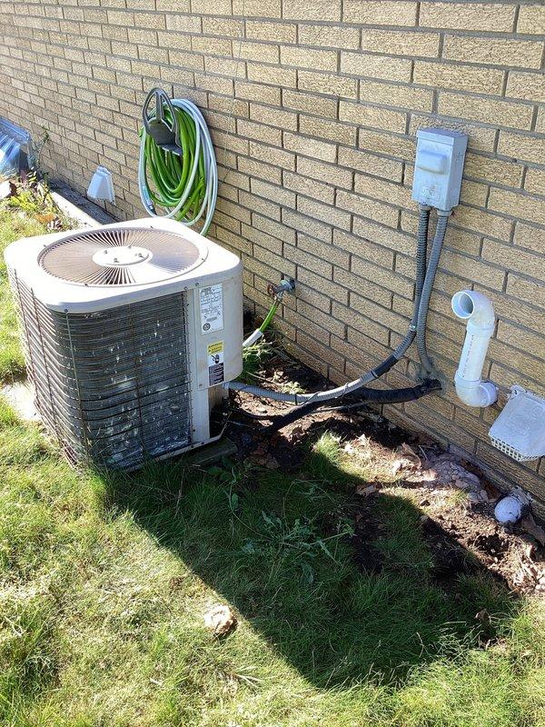 Responded to no-cool complaint with outdoor unit not running and warm air from vents. Upon arrival, found white cylindrical condenser unit positioned against exterior brick wall with airflow present at indoor registers and no ice buildup at evaporator coil. Performed visual inspection of outdoor unit, verified electrical disconnect box and refrigerant line connections, and documented condenser placement and surrounding installation components including PVC condensate drain and refrigerant line routing along foundation. System age unknown but reported as potentially under 10 years old.