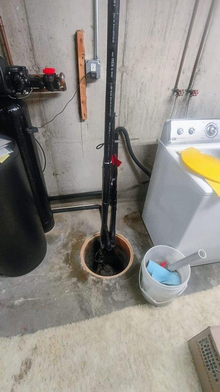 Responded to emergency call for malfunctioning sump pump in 1970s-era home basement. Upon arrival, assessed reported symptoms of unusual thumping noise and visible smoke emanating from pump unit prior to customer disconnecting power. Conducted visual inspection of sump pit area located near laundry facilities, noting moisture accumulation and pump assembly condition. Documented existing installation configuration including discharge piping, electrical connections, and pump housing with significant water exposure. Evaluated gray mechanical pump unit with multiple mounting bolts and observed wet concrete floor conditions around basin. Provided estimate for complete sump pump replacement to restore proper basement water removal functionality and prevent potential flooding issues.