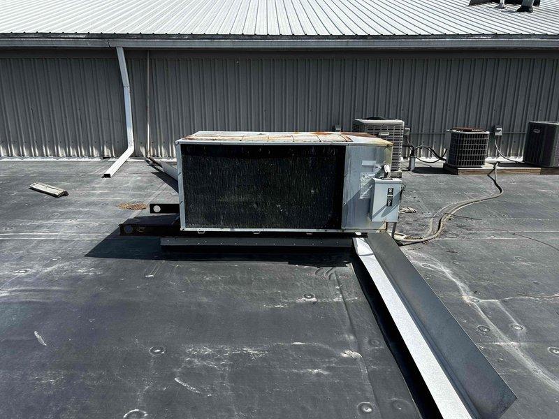 Conducted rooftop HVAC assessment for two barely-functioning air conditioning systems on commercial property. Evaluated multiple rooftop package units installed on elevated platform with rubber membrane roofing, documenting existing equipment conditions including cylindrical condenser unit with cage housing and rectangular rooftop AC units mounted on metal curbs. Photographed current system configurations, electrical disconnects, conduit routing, and mounting platforms to prepare comprehensive replacement estimate for dual AC system installation.