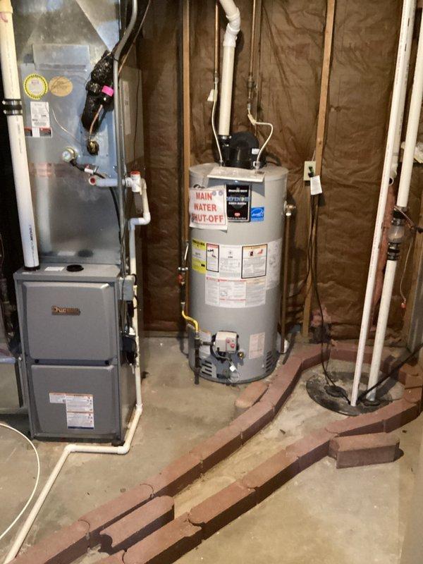 Responded to service call for reported water heater leak in basement with visible pudding around unit. Upon arrival, conducted thorough inspection of white cylindrical tanked water heater and surrounding plumbing connections. Examined all visible piping including copper supply lines, temperature and pressure relief valve, drain valve, and discharge tube for signs of active leakage or moisture accumulation. Inspected tank exterior for corrosion, rust staining, or structural compromise. Checked floor area beneath and around water heater to identify source and extent of water accumulation on concrete floor. Verified unit was currently operational and producing hot water as reported. Assessed age and condition of installation based on manufacturer labels and specifications visible on tank exterior. Evaluated all connection points, fittings, and valves for potential leak sources. Documented findings with photographic evidence showing mechanical room layout, water heater positioning relative