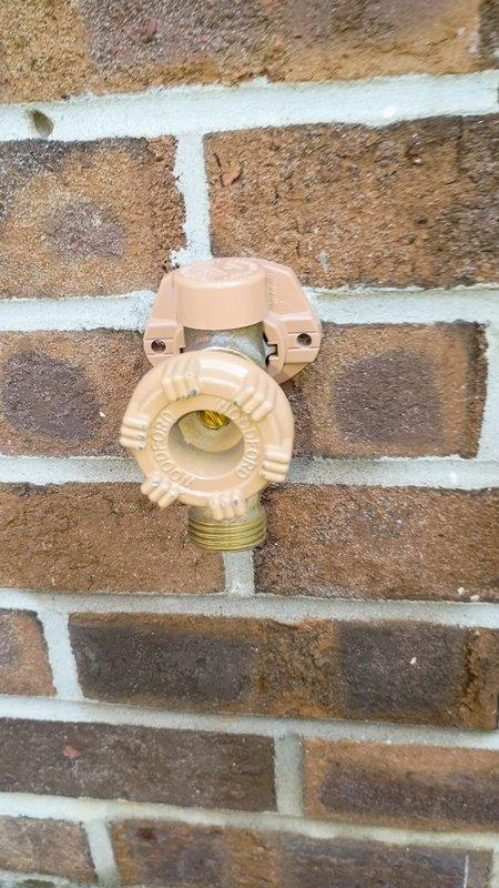 Responded to report of leaking outdoor water spigot mounted on exterior brick wall. Upon inspection, identified beige polymer hose bib with brass threaded outlet showing signs of water seepage. Evaluated fixture mounting, valve body integrity, and connection points to determine source of leak. Documented existing conditions of spigot assembly and surrounding area including landscaping rock bed and drainage components at base of wall.