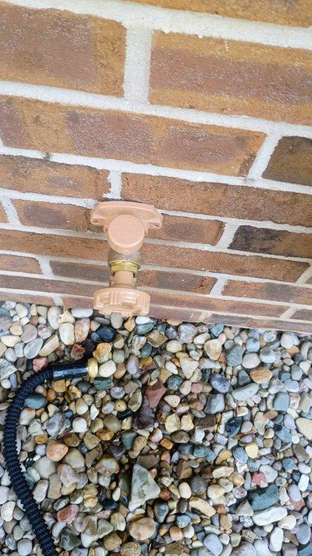 Responded to report of leaking outdoor water spigot mounted on exterior brick wall. Upon inspection, identified beige polymer hose bib with brass threaded outlet showing signs of water seepage. Evaluated fixture mounting, valve body integrity, and connection points to determine source of leak. Documented existing conditions of spigot assembly and surrounding area including landscaping rock bed and drainage components at base of wall.