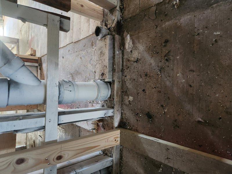 Continued plumbing rough-in work on active construction project, installing PVC drainage piping with proper support brackets and elbow fittings in basement area with new wooden framing. Exterior drainage line routed through deck structure using white PVC piping to integrate with building's plumbing system.
