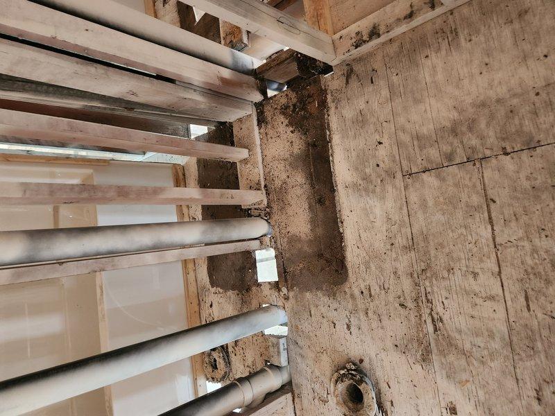 Continued plumbing rough-in work for central heating system installation. Installed and routed multiple horizontal PVC supply and drain lines through wooden framing members at various elevations, ensuring proper spacing and support. Set HVAC condensate drain pan with integrated drain fitting within metal framework structure above mechanical room area, positioning unit to accommodate evaporator coil assembly with exposed copper tubing.