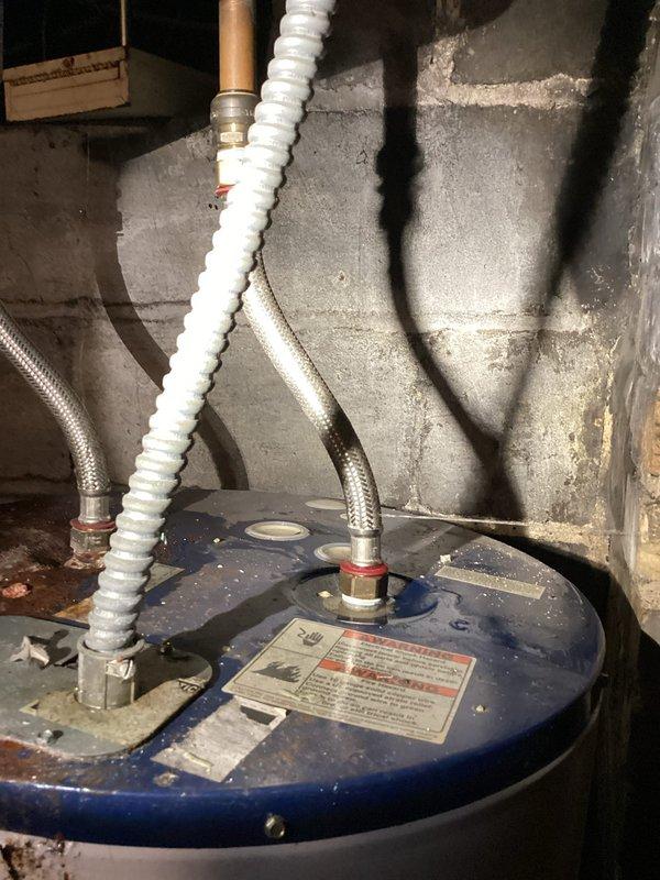 Responded to emergency water heater leak with main water supply already shut off upon arrival. Inspected aging tanked water heater and identified failed flexible supply hose connection at top of unit spraying water prior to shutoff. Observed significant corrosion and rust staining on tank top and body indicating advanced age and deterioration of unit. Documented existing conditions showing corroded black top surface, rust stains on white tank exterior, and compromised flexible connector fittings. Due to extensive corrosion, age-related deterioration, and active leak from supply line connection, recommended full water heater replacement rather than repair of failed component on compromised unit.