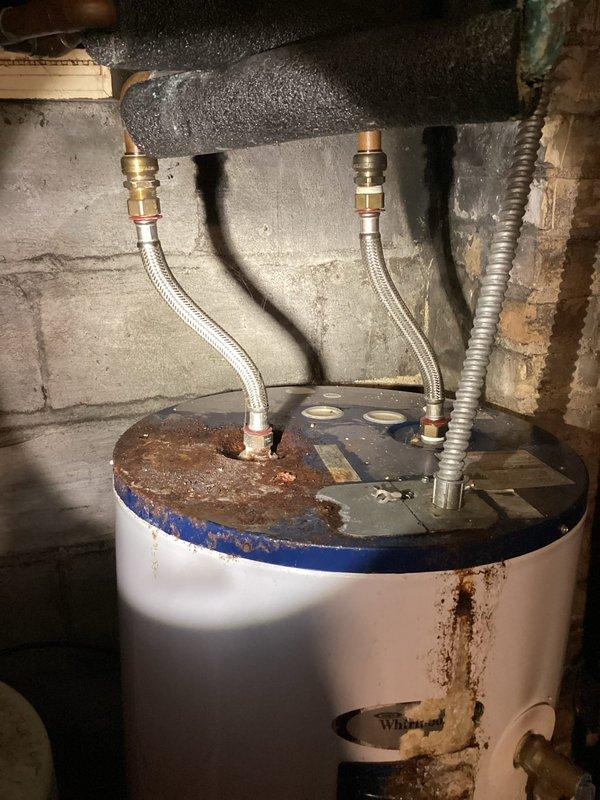 Responded to emergency water heater leak with main water supply already shut off upon arrival. Inspected aging tanked water heater and identified failed flexible supply hose connection at top of unit spraying water prior to shutoff. Observed significant corrosion and rust staining on tank top and body indicating advanced age and deterioration of unit. Documented existing conditions showing corroded black top surface, rust stains on white tank exterior, and compromised flexible connector fittings. Due to extensive corrosion, age-related deterioration, and active leak from supply line connection, recommended full water heater replacement rather than repair of failed component on compromised unit.
