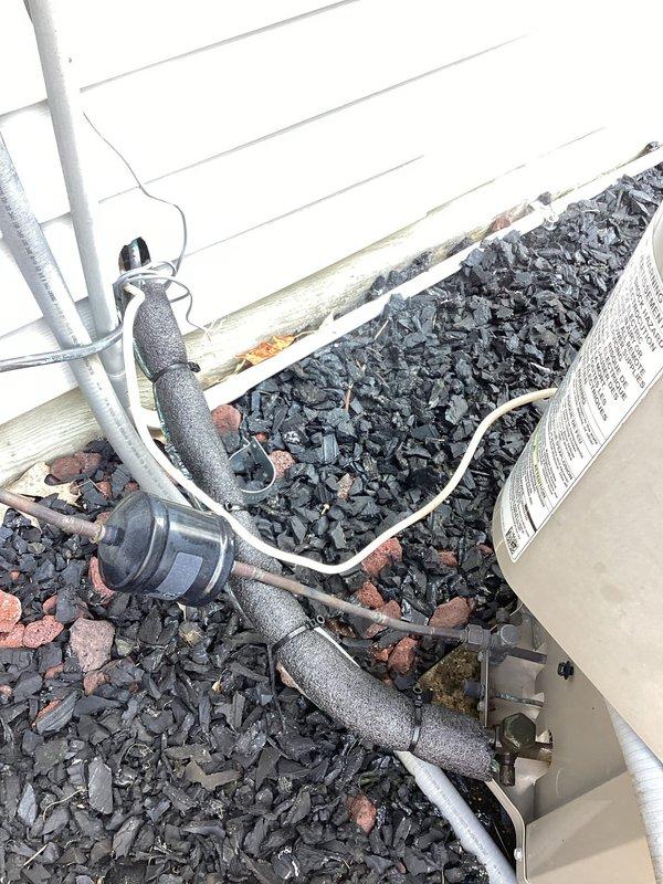 # HVAC Maintenance - Planned Single Maintenance

Completed comprehensive air conditioning maintenance inspection on residential outdoor condensing unit. Performed visual assessment of refrigerant line set, noting weathered gray foam insulation on suction line with signs of UV degradation and age-related deterioration. Inspected copper refrigerant lines for proper routing and support, observing surface oxidation consistent with normal outdoor exposure. 

Examined electrical conduit and wiring connections running to condensing unit, verifying secure mounting to exterior wall. Assessed white PVC condensate drain line for proper pitch and drainage functionality. Verified unit positioning on gravel pad with adequate clearance from building structure. Inspected manufacturer label and unit specifications for reference during maintenance procedures. All refrigerant lines properly insulated and secured with appropriate mounting brackets. System installation shows typical aging characteristics b