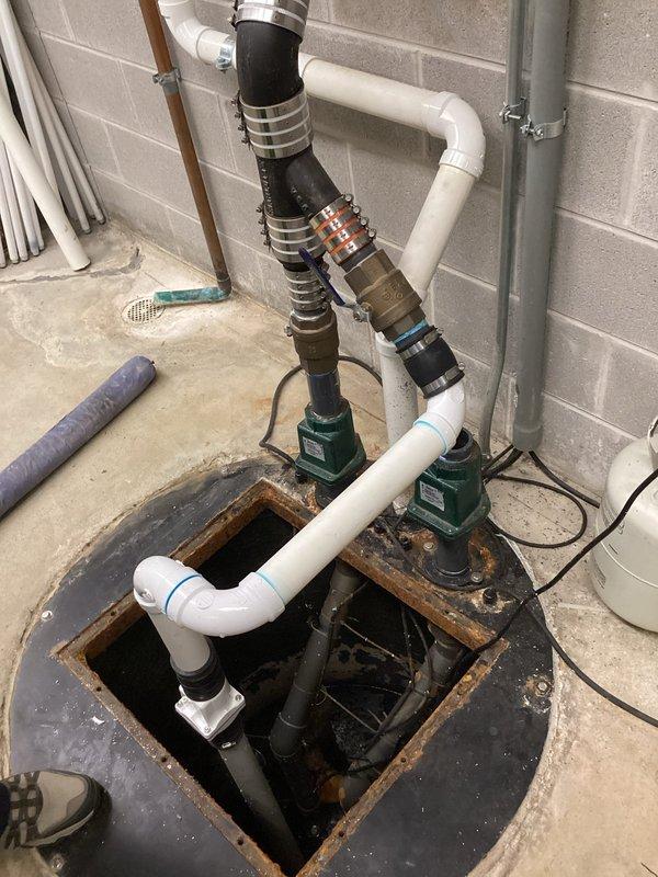 Conducted on-site assessment of existing dual sump pump system in residential basement to provide replacement quote. Inspected two green submersible pumps installed in single basin with white PVC discharge piping and black corrugated flexible drainage hose. Observed significant rust and corrosion on metal pit cover, mineral deposits and wear throughout system, and aged plumbing connections. Documented current configuration including multiple drain lines feeding into basin, vertical discharge piping with compression fittings, and overall system condition. Photographed installation setup and discussed replacement options for both pumps with property manager.