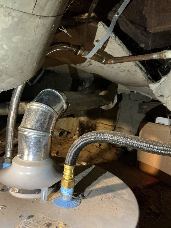 Completed inspection and service of tanked water heater installation in basement crawl space. Assessed existing plumbing connections including braided stainless steel flex lines, PVC drainage system, and galvanized ductwork. Documented condition of water heater connections, including metallic elbow pipe with oxidation, brass fittings at junction points, and flex line connections with blue-colored fittings. Verified proper installation of metal strapping securing pipes to structural joists and confirmed all connections were secure and functioning properly.