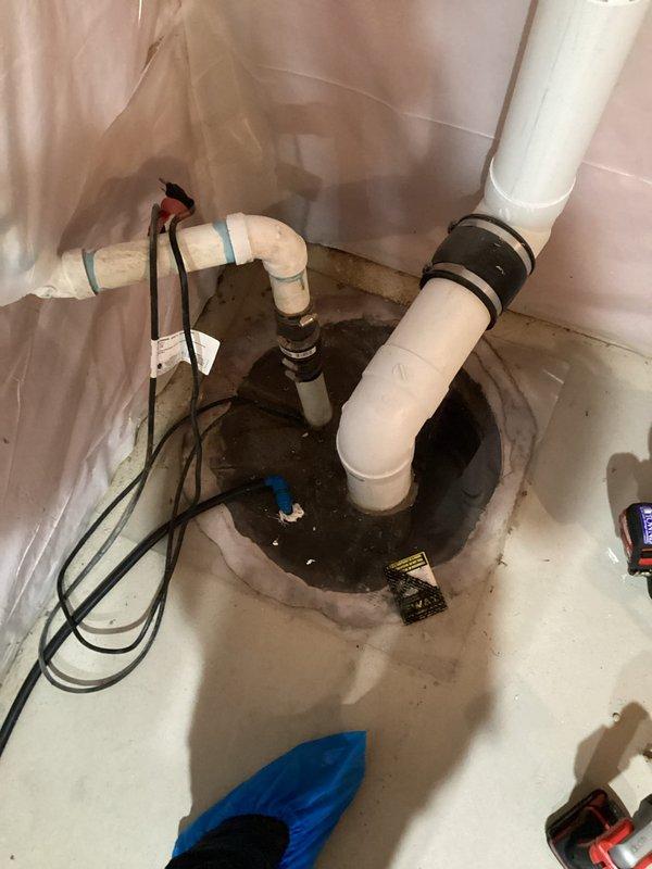 Responded to sump pump malfunction in residential basement that had been experiencing issues for approximately 10 days. Upon arrival, accessed the work area and conducted thorough inspection of the existing sump pump system installed in a concrete pit. Found significant mineral deposits, staining, and debris accumulation throughout the basin, indicating extended operation in challenging water conditions. Examined the pump mechanism, discharge piping configuration with multiple PVC connections, and electrical components. Documented the condition of the circular concrete sump basin showing wear consistent with a 20-year-old installation, including visible moisture damage, concrete deterioration around the perimeter, and standing water at the basin bottom. Assessed the pump assembly, hose clamps, and discharge connections for proper operation and potential failure points. Protected surrounding work area with plastic sheeting during inspection. Photographed the complete installation from m