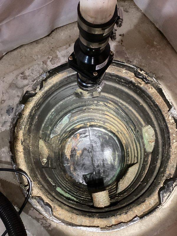 Responded to sump pump malfunction in residential basement that had been experiencing issues for approximately 10 days. Upon arrival, accessed the work area and conducted thorough inspection of the existing sump pump system installed in a concrete pit. Found significant mineral deposits, staining, and debris accumulation throughout the basin, indicating extended operation in challenging water conditions. Examined the pump mechanism, discharge piping configuration with multiple PVC connections, and electrical components. Documented the condition of the circular concrete sump basin showing wear consistent with a 20-year-old installation, including visible moisture damage, concrete deterioration around the perimeter, and standing water at the basin bottom. Assessed the pump assembly, hose clamps, and discharge connections for proper operation and potential failure points. Protected surrounding work area with plastic sheeting during inspection. Photographed the complete installation from m