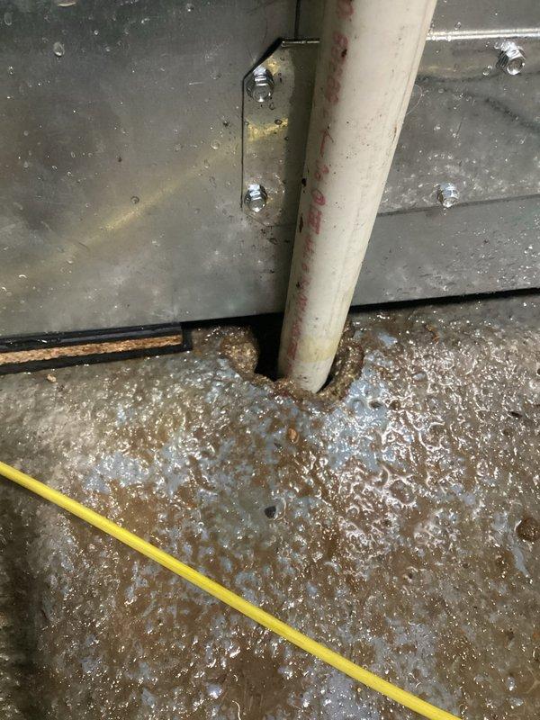Responded to water leak complaint in basement originating from dehumidifier system. Upon arrival, found significant standing water accumulation with rust-colored discoloration pooling around base of HVAC equipment and metal ductwork assembly. Inspected white PVC condensate drain line running vertically along galvanized metal panels, noting visible moisture and discoloration on pipe surface. Examined connection points, elbow joints, and horizontal drain sections for potential leak sources. Assessed air handler unit with dark gray metal panel and associated drain piping configuration. Documented water damage to concrete floor with extensive pooling around equipment base, indicating ongoing drainage issue. Photographed affected areas showing water contact on metal surfaces, electrical components, and floor substrate for service records.