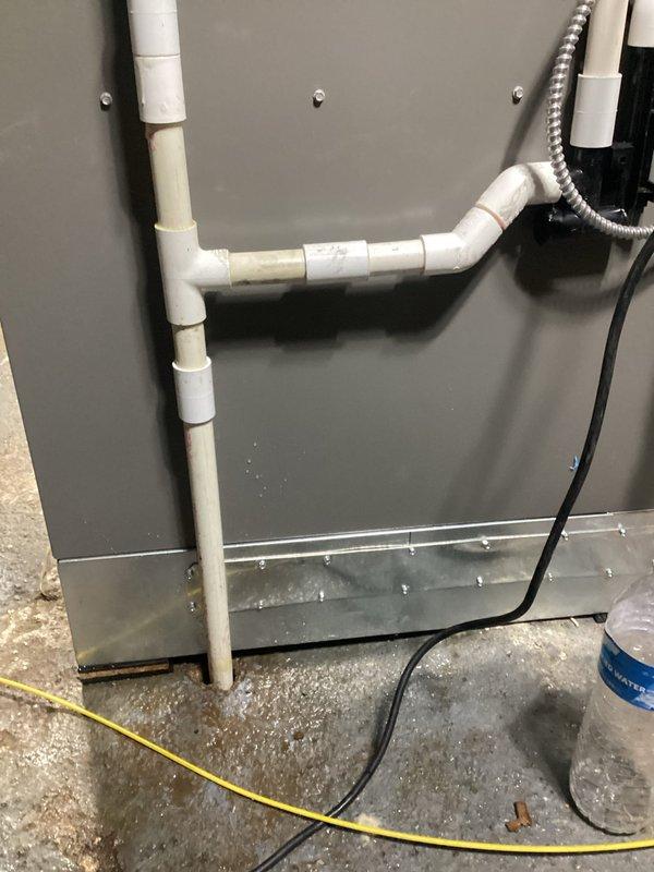 Responded to water leak complaint in basement originating from dehumidifier system. Upon arrival, found significant standing water accumulation with rust-colored discoloration pooling around base of HVAC equipment and metal ductwork assembly. Inspected white PVC condensate drain line running vertically along galvanized metal panels, noting visible moisture and discoloration on pipe surface. Examined connection points, elbow joints, and horizontal drain sections for potential leak sources. Assessed air handler unit with dark gray metal panel and associated drain piping configuration. Documented water damage to concrete floor with extensive pooling around equipment base, indicating ongoing drainage issue. Photographed affected areas showing water contact on metal surfaces, electrical components, and floor substrate for service records.