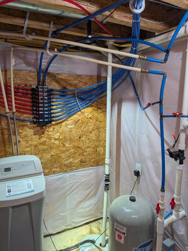 Responded to service call for non-operational tanked water heater with no status light indication and complete loss of hot water. Upon arrival, found unit was over 10 years old with no visible leaks present. Inspected basement utility area containing water heater, water softener system, expansion tank, and extensive PEX plumbing distribution manifold. Performed diagnostic evaluation of water heater electrical components, gas supply connections, and control systems. Assessed status indicator functionality and verified power supply to unit. Examined associated plumbing connections including hot and cold water lines, pressure relief valve, and expansion tank operation. Documented existing plumbing infrastructure including blue PEX cold water lines, red PEX hot water distribution, and white PVC drainage systems throughout unfinished basement space. Reviewed overall system configuration with customer and provided recommendations for repair versus replacement options given age and condition 