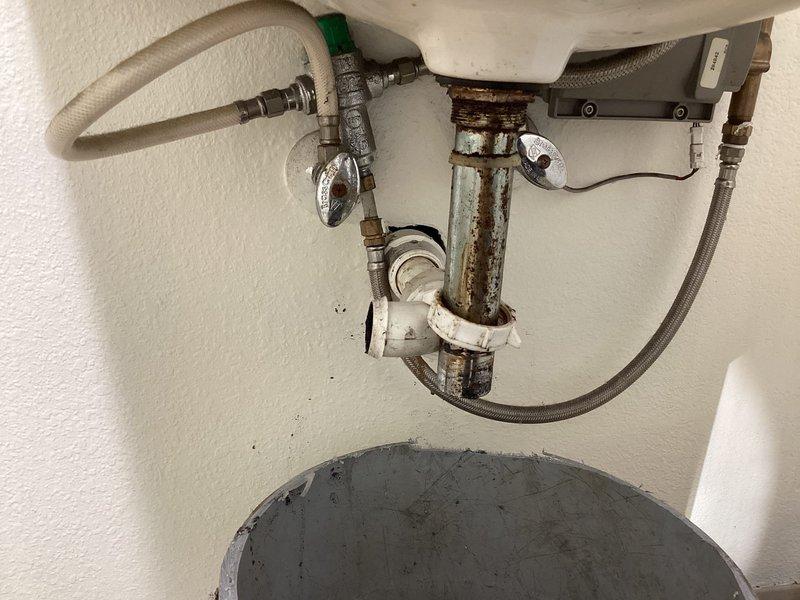 Responded to urgent call for slow-draining bathroom sink on shop floor. Initial attempt to clear blockage by removing and cleaning P-trap was unsuccessful in resolving drainage issue. Conducted thorough inspection of drain assembly and accessible plumbing components beneath sink, including PVC drain piping, trap configuration, and supply line connections. Documented existing conditions of drainage system showing age-related discoloration and mineral deposits on fixtures. Inspected associated plumbing infrastructure including corroded metal pipes, valves, and water heater connections displaying significant rust and oxidation. Photographed all plumbing components for service records. Blockage determined to be located beyond P-trap in main drain line, requiring additional drain cleaning equipment or methods to fully resolve slow drainage condition.