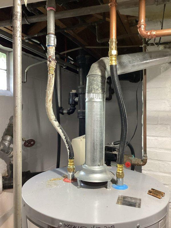 Responded to service call for 7-year-old tanked water heater with reported water leakage from bottom of unit and inoperative pilot light with no status light indication. Upon arrival, performed comprehensive diagnostic inspection of gas control valve, burner assembly, and tank integrity. Removed access panel to examine internal components including gas control valve with status light indicator, flexible gas line connector, and burner assembly with brass fittings and copper connections. Inspected expansion tank mounted on top of unit with flexible braided hoses and brass fittings, along with associated copper piping and PVC connections running to ceiling joists. Identified active leak originating from lower tank section and confirmed pilot light failure with non-functional status indicator on gas control valve. Assessed overall system configuration including plumbing connections, gas supply line, and venting arrangement. Documented conditions through photographic evidence showing compro