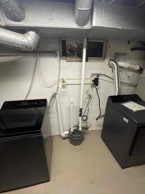 Installed above-ground sump pump system in basement utility room between laundry appliances. System includes white PVC discharge piping assembly with black cylindrical basin positioned at floor level, with vertical piping extending upward to connect with existing drainage infrastructure. Installation completed in unfinished basement area with proper routing of discharge lines through exposed joist space to ensure effective water removal and code-compliant operation.