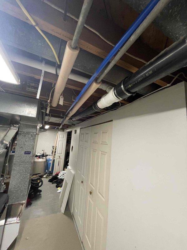 Installed above-ground sump pump system in basement utility room between laundry appliances. System includes white PVC discharge piping assembly with black cylindrical basin positioned at floor level, with vertical piping extending upward to connect with existing drainage infrastructure. Installation completed in unfinished basement area with proper routing of discharge lines through exposed joist space to ensure effective water removal and code-compliant operation.