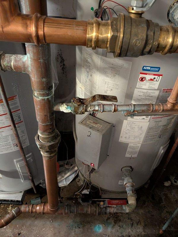Responded to service call for non-operational tanked water heater system with reported leak. Upon inspection, identified active leak originating from cold water supply line connection to water heater rather than tank itself. System consists of dual white cylindrical water heaters with 200,000 BTU combined capacity, approximately one year old. 

Observed extensive corrosion and mineral deposits on multiple pipe fittings and connections throughout the installation, including heavy rust on horizontal supply lines and green oxidation (verdigris) at copper joints. The compromised water line connection was causing system shutdown and water damage to surrounding area. Brass valve assembly at top of system showed signs of wear with associated electrical wiring connections intact.

Drained water heater tanks completely to facilitate safe repair of leaking connection point. Inspected expansion tank, pressure relief valves, and associated copper and steel piping for additional compromise. Noted s