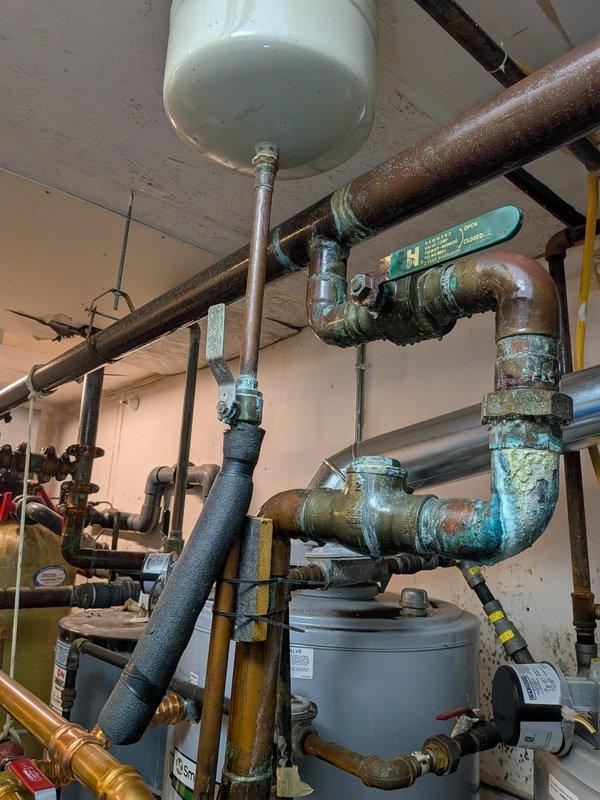 Responded to service call for non-operational tanked water heater system with reported leak. Upon inspection, identified active leak originating from cold water supply line connection to water heater rather than tank itself. System consists of dual white cylindrical water heaters with 200,000 BTU combined capacity, approximately one year old. 

Observed extensive corrosion and mineral deposits on multiple pipe fittings and connections throughout the installation, including heavy rust on horizontal supply lines and green oxidation (verdigris) at copper joints. The compromised water line connection was causing system shutdown and water damage to surrounding area. Brass valve assembly at top of system showed signs of wear with associated electrical wiring connections intact.

Drained water heater tanks completely to facilitate safe repair of leaking connection point. Inspected expansion tank, pressure relief valves, and associated copper and steel piping for additional compromise. Noted s